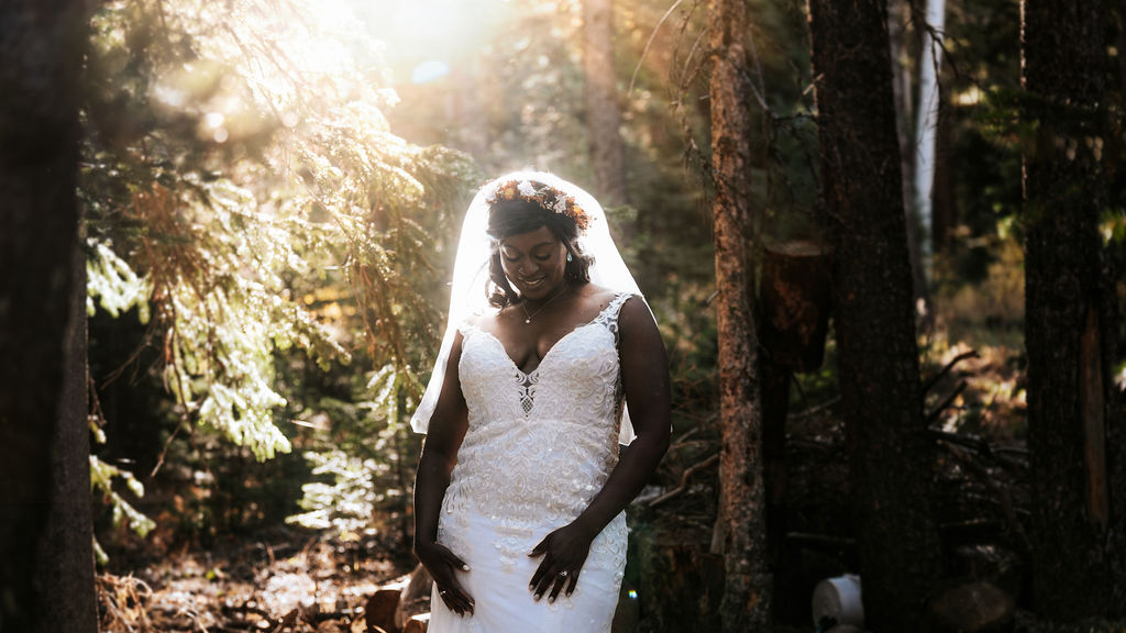 A bride in a lace dress smiles while looking down and standing in a forest at sunset during her crested butte elopement