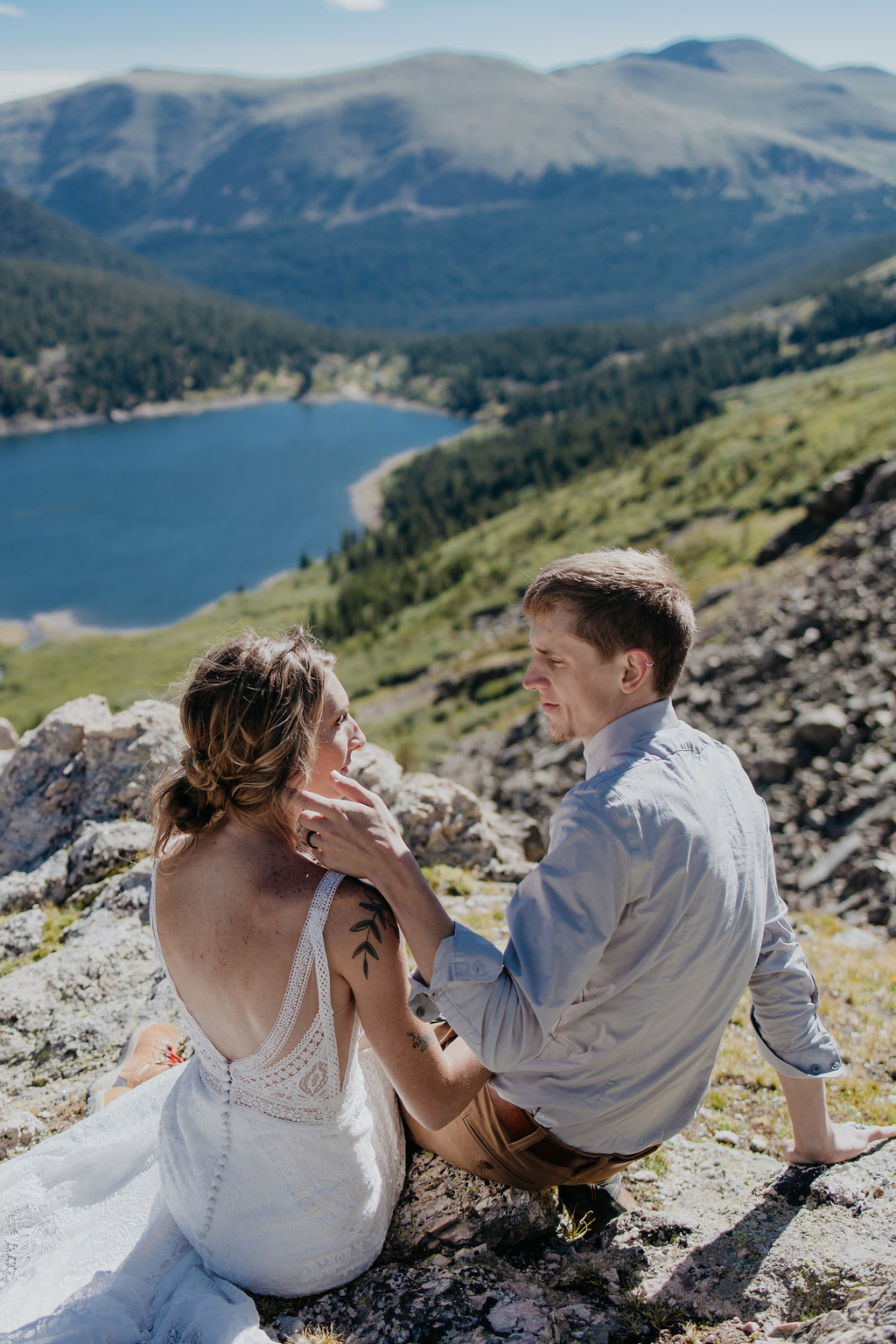 Newlyweds sit on a rock overlooking a mountain lake in a valley below looking at each other