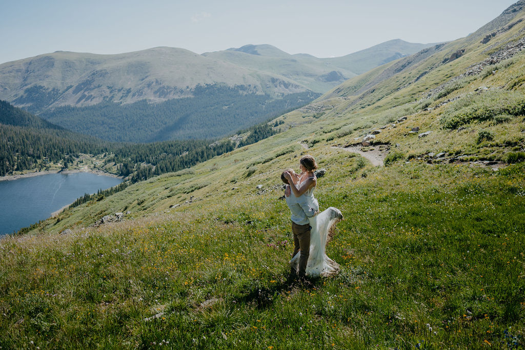 A groom lifts his bride on a hillside overlooking mountains and a lake during their denver elopement