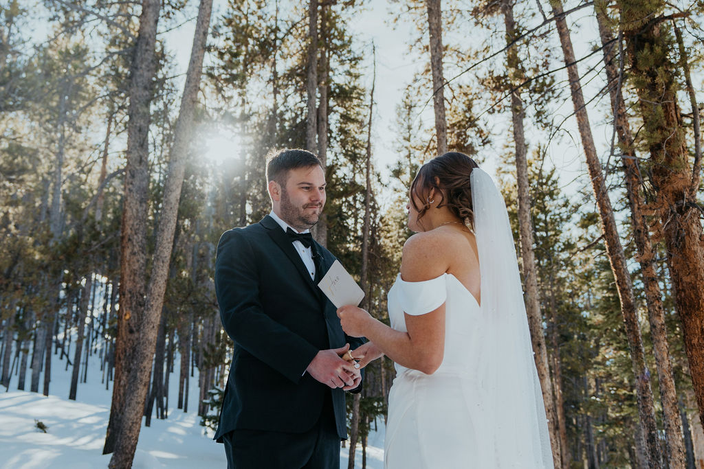 A bride reads her vows to her husband in a black suit in a snow covered forest