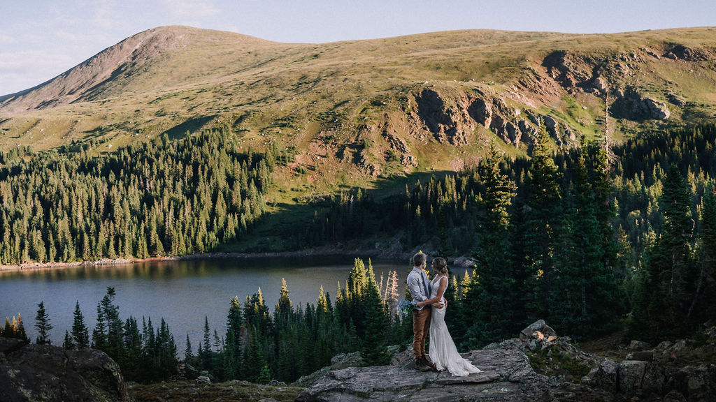 Newlyweds stand on a rock overlooking a mountain lake hugging during their boulder elopement