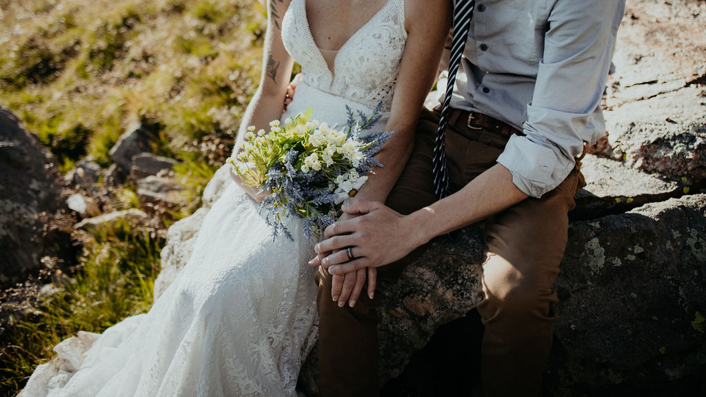 Newlyweds sit close holding hands on a mountain rock during their boulder elopement