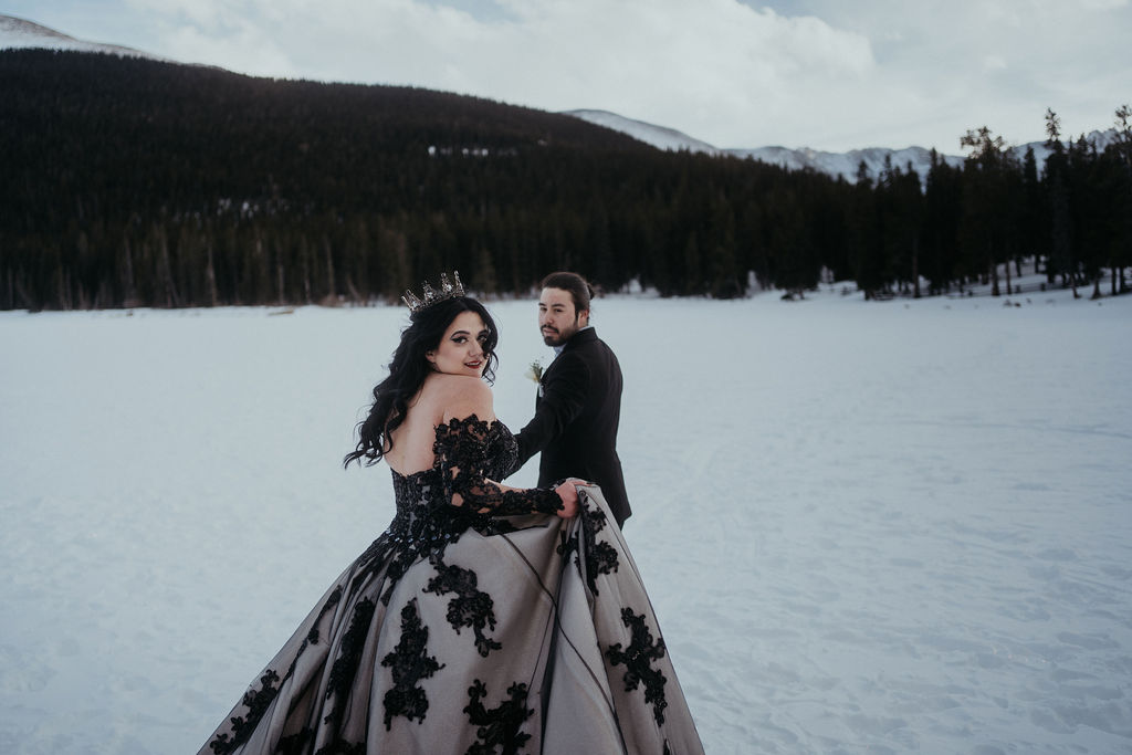 A groom in a black suit leads his bride by the hand through a snowy field in her black lace dress